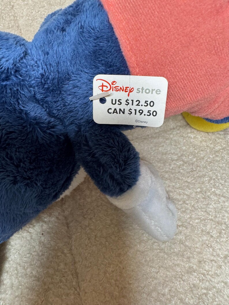 May include: A soft toy in blue, pink, and white, with a Disney Store tag. The tag features the Disney Store logo and the text "US $12.50" and "CAN $19.50". The toy is made of a fluffy material.