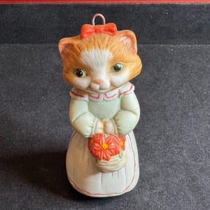 May include: A ceramic figurine of a cat with orange and white fur, wearing a light green dress and holding a basket of red flowers. The cat has a red bow on its head and a metal loop for hanging.