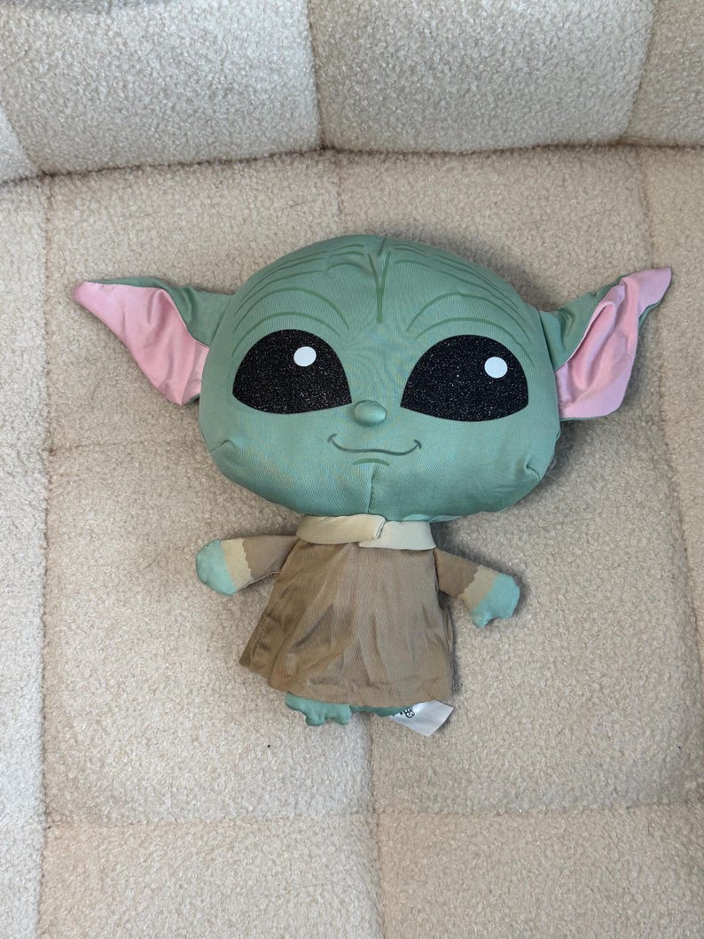 May include: A plush toy of a character with large ears, large black eyes, and a small smile. The character has a green head, pink ears, and wears a tan robe. The toy is approximately 12 inches tall.