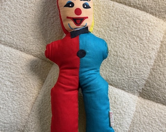 May include: A vintage clown doll with a red and blue body, a yellow and red hat, and a smiling face. The doll has a black bow tie and a black button on the front. The doll is made of fabric and is likely a collectible item.
