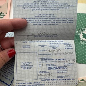 May include: A Cabbage Patch Kids passport document with a blue patterned cover. The interior page features printed text in English and French, a signature line for a parent, and handwritten details including the name Martie Fayette Holleran.