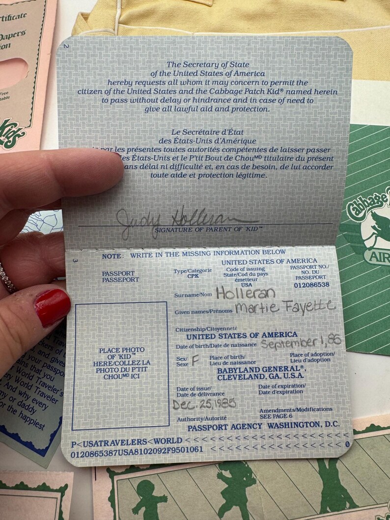 May include: A Cabbage Patch Kids passport document with a blue patterned cover. The interior page features printed text in English and French, a signature line for a parent, and handwritten details including the name Martie Fayette Holleran.