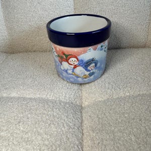May include: A ceramic planter with a dark blue rim and a white interior. The exterior features a winter scene with two snowmen sledding down a snowy hill. The snowmen are wearing hats and scarves, and the background has snowflakes.