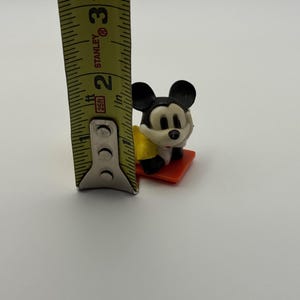 May include: A small Mickey Mouse figurine, approximately 1.5 inches tall, is positioned on a red base. The figure is black, white, and yellow. A yellow and black measuring tape is in the frame.