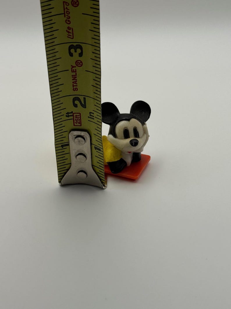 May include: A small Mickey Mouse figurine, approximately 1.5 inches tall, is positioned on a red base. The figure is black, white, and yellow. A yellow and black measuring tape is in the frame.