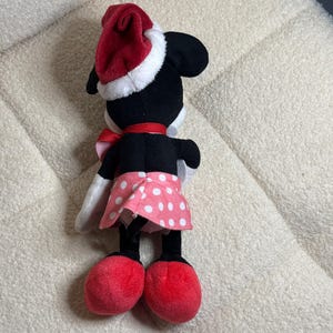 Op de afbeelding: Een pluche Minnie Mouse pop, gezien van achteren, met een rode kerstmuts met witte rand, een rode strik en een roze rok met stippen. De pop heeft zwarte oren, een zwart lichaam en rode schoenen. Een feestelijk kerstartikel.