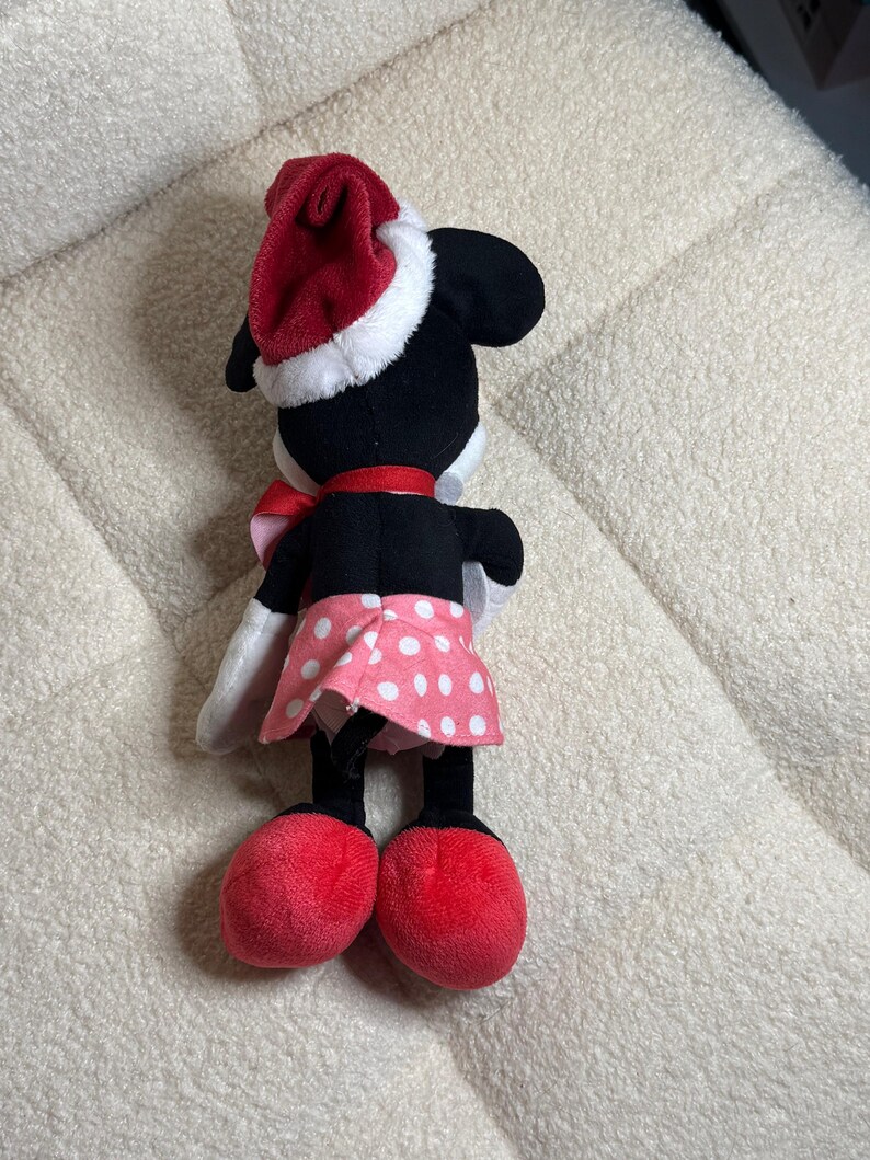 Op de afbeelding: Een pluche Minnie Mouse pop, gezien van achteren, met een rode kerstmuts met witte rand, een rode strik en een roze rok met stippen. De pop heeft zwarte oren, een zwart lichaam en rode schoenen. Een feestelijk kerstartikel.
