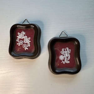 May include: Two small, framed wall hangings with dark brown frames. Each frame contains a burgundy background and a white lace design. One features two birds and flowers, the other a butterfly. Both have metal triangle hangers.