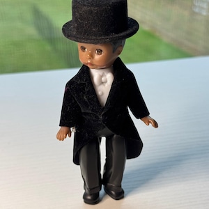 May include: A small doll dressed in a black tuxedo with a matching top hat. The doll has dark skin and is wearing a white bow tie and shirt. The doll's black pants and shoes complete the formal attire.