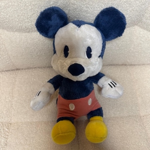 May include: A soft toy of Mickey Mouse, featuring navy blue ears, a white face, and a black nose. He is dressed in red shorts with white buttons, yellow shoes, and white gloves with black stripes.