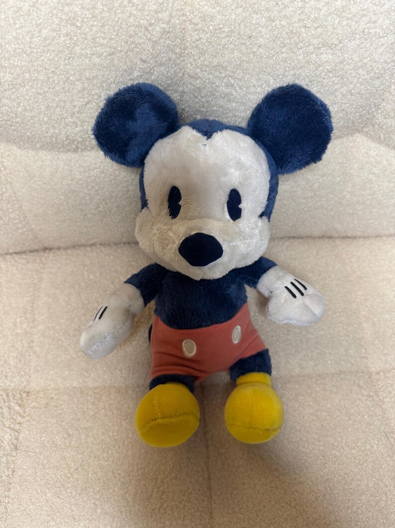May include: A soft toy of Mickey Mouse, featuring navy blue ears, a white face, and a black nose. He is dressed in red shorts with white buttons, yellow shoes, and white gloves with black stripes.