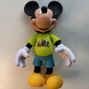 May include: A Mickey Mouse figurine with black ears, a tan face, and a black nose. He wears a yellow shirt with a graphic, blue shorts, white gloves, and yellow shoes. The figurine is standing upright.