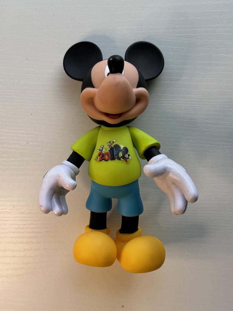 May include: A Mickey Mouse figurine with black ears, a tan face, and a black nose. He wears a yellow shirt with a graphic, blue shorts, white gloves, and yellow shoes. The figurine is standing upright.