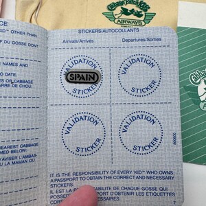May include: A Cabbage Patch Kids passport booklet with blue text and grid patterns, featuring four circular outlines labelled Validation Sticker. One silver sticker with the word SPAIN is placed over the top-left circle. A yellow bag is visible in the background.