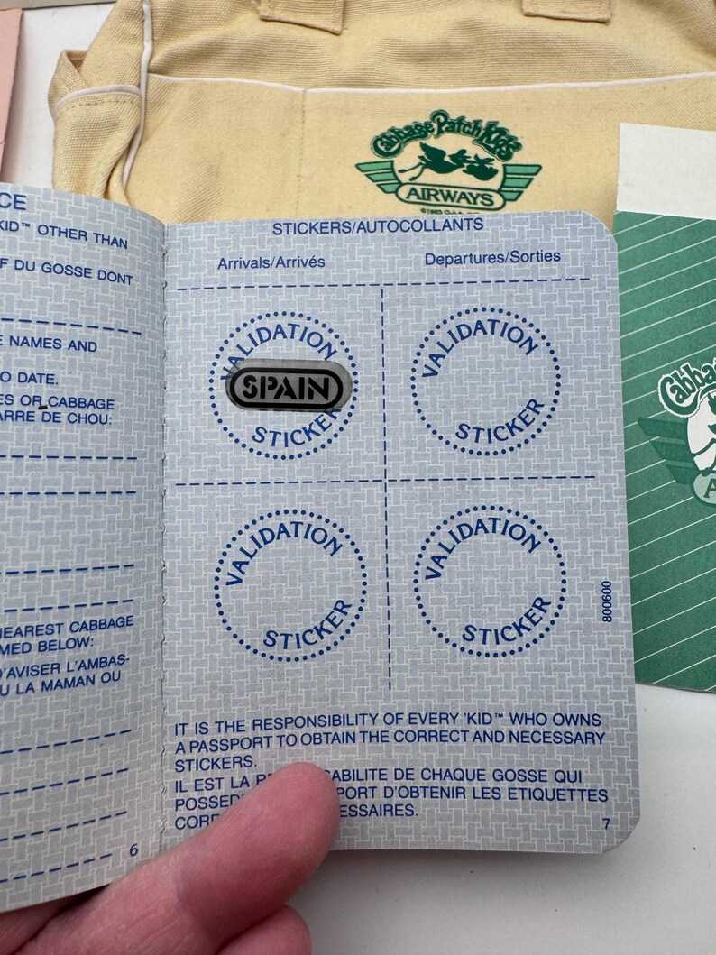 May include: A Cabbage Patch Kids passport booklet with blue text and grid patterns, featuring four circular outlines labelled Validation Sticker. One silver sticker with the word SPAIN is placed over the top-left circle. A yellow bag is visible in the background.