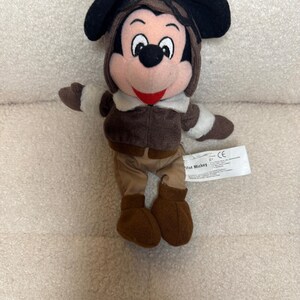 K&ouml;nnte beinhalten: Ein Stofftier von Mickey Mouse in einem braunen Fliegeroutfit, einschlie&szlig;lich einer M&uuml;tze mit Ohrenklappen, einer Jacke, einer Hose und Stiefeln. Das Spielzeug hat schwarze Ohren, ein rosa Gesicht und ein rotes L&auml;cheln. Ein wei&szlig;es Etikett ist angebracht.