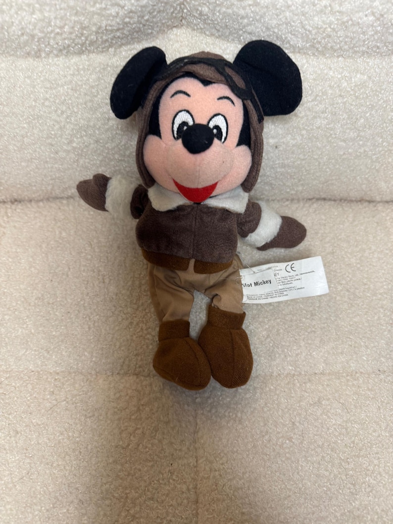 K&ouml;nnte beinhalten: Ein Stofftier von Mickey Mouse in einem braunen Fliegeroutfit, einschlie&szlig;lich einer M&uuml;tze mit Ohrenklappen, einer Jacke, einer Hose und Stiefeln. Das Spielzeug hat schwarze Ohren, ein rosa Gesicht und ein rotes L&auml;cheln. Ein wei&szlig;es Etikett ist angebracht.
