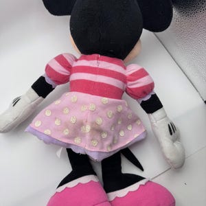May include: A plush Minnie Mouse doll, viewed from the back, with black ears and head, wearing a pink and white striped top and a pink polka-dot skirt. The doll has white gloves, black arms and legs, and pink shoes.