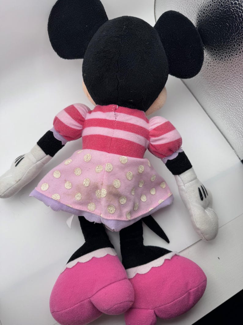 May include: A plush Minnie Mouse doll, viewed from the back, with black ears and head, wearing a pink and white striped top and a pink polka-dot skirt. The doll has white gloves, black arms and legs, and pink shoes.