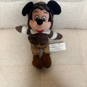 K&ouml;nnte beinhalten: Ein Stofftier von Mickey Mouse als Pilot. Die Figur tr&auml;gt einen braunen Fliegerhut mit Brille, eine braune Jacke mit wei&szlig;em Besatz und eine braune Hose und Stiefel. Mickey hat schwarze Ohren, ein rosa Gesicht und ein rotes L&auml;cheln.