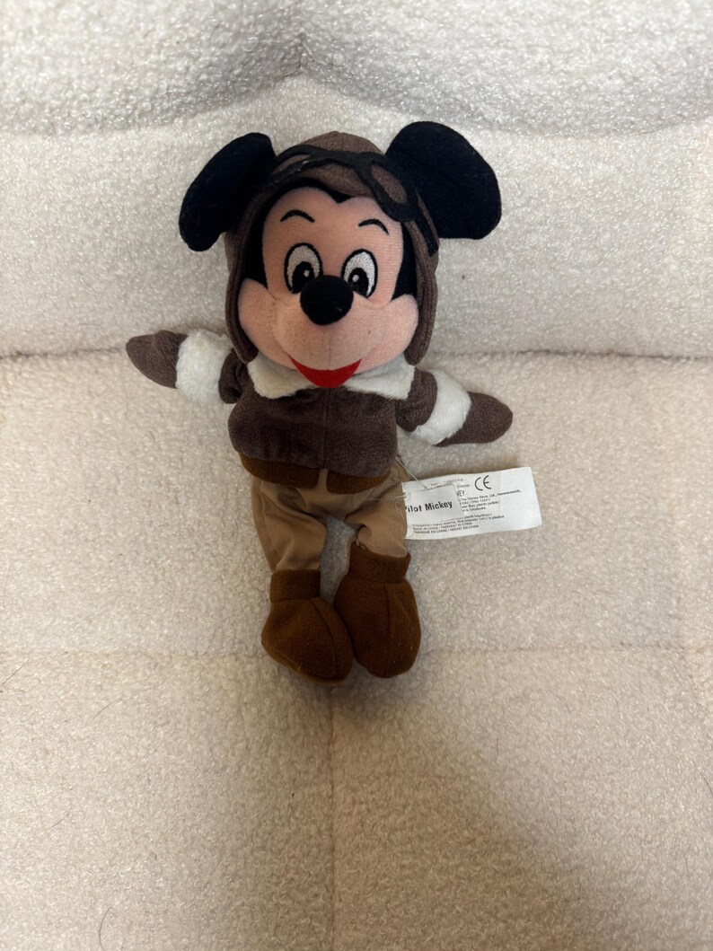 K&ouml;nnte beinhalten: Ein Stofftier von Mickey Mouse als Pilot. Die Figur tr&auml;gt einen braunen Fliegerhut mit Brille, eine braune Jacke mit wei&szlig;em Besatz und eine braune Hose und Stiefel. Mickey hat schwarze Ohren, ein rosa Gesicht und ein rotes L&auml;cheln.
