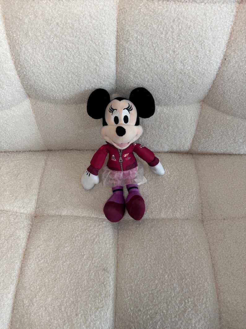 Puede incluir: Un mu&ntilde;eco de peluche de Minnie Mouse, con orejas negras, cara blanca y una chaqueta roja con cremallera. Lleva una falda rosa, calcetines morados y zapatos rojo oscuro. La mu&ntilde;eca est&aacute; colocada sobre una superficie texturizada de color blanco roto.