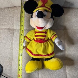 May include: A plush Minnie Mouse doll wearing a yellow marching band uniform with red accents. The doll has black ears, a yellow hat, and measures approximately 18 inches in height.
