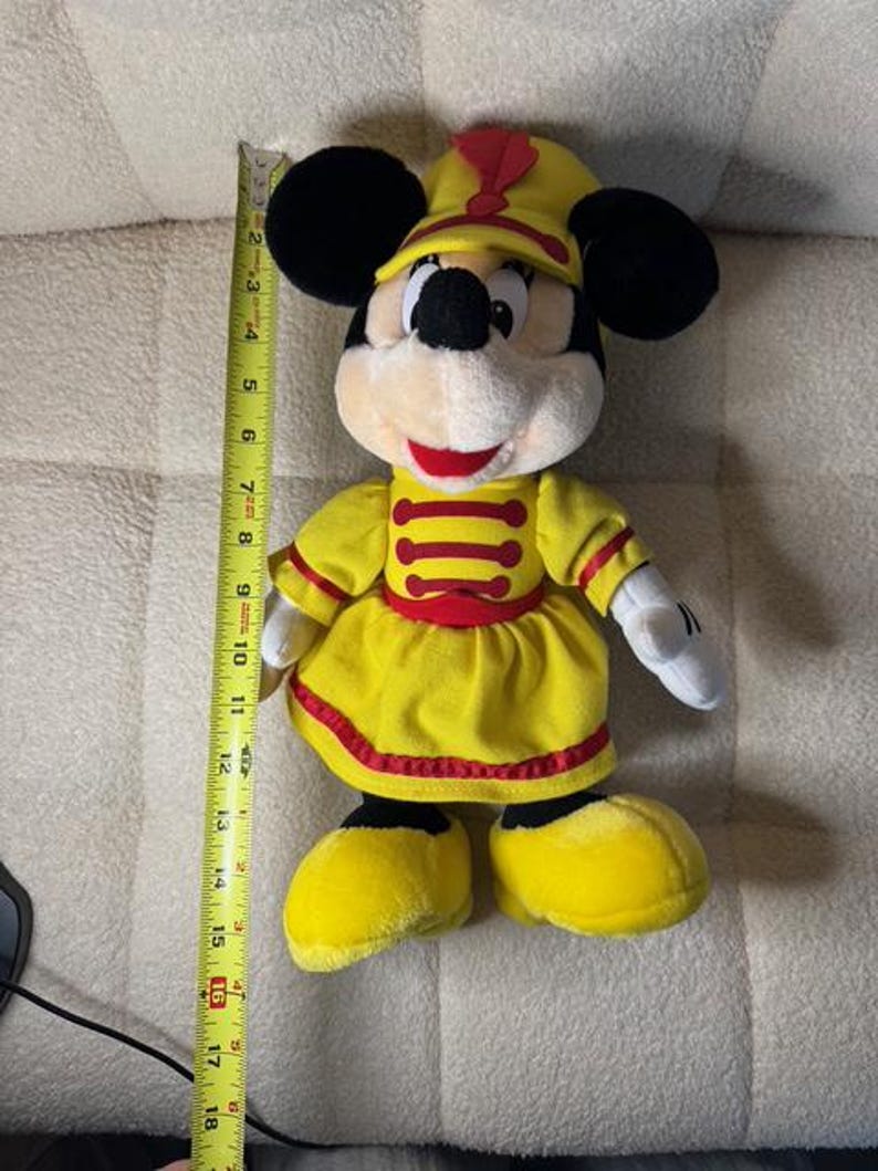 May include: A plush Minnie Mouse doll wearing a yellow marching band uniform with red accents. The doll has black ears, a yellow hat, and measures approximately 18 inches in height.