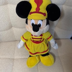 May include: A plush Minnie Mouse doll wearing a yellow marching band uniform with a red accent. The doll has black ears, a black nose, and white gloves. The hat has a red feather. The doll is standing on a cream-colored surface.