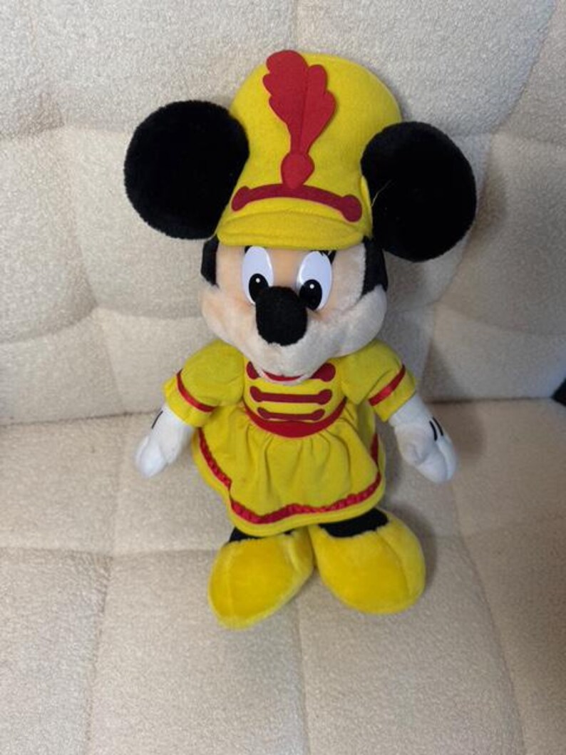 May include: A plush Minnie Mouse doll wearing a yellow marching band uniform with a red accent. The doll has black ears, a black nose, and white gloves. The hat has a red feather. The doll is standing on a cream-colored surface.