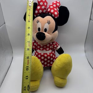 May include: A plush Minnie Mouse doll, approximately 12 inches tall, with a red and white polka dot dress and bow. The doll has black ears, a black nose, and yellow feet. The doll is sitting upright.