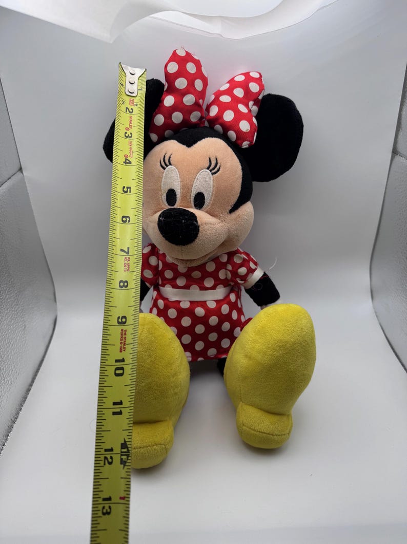 May include: A plush Minnie Mouse doll, approximately 12 inches tall, with a red and white polka dot dress and bow. The doll has black ears, a black nose, and yellow feet. The doll is sitting upright.