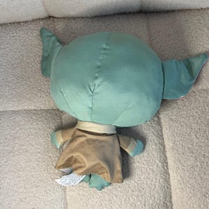 May include: A plush doll of a character with large ears, a green head, and a tan robe. The doll has small arms and feet with blue accents. The doll is lying on a light-colored, textured surface.