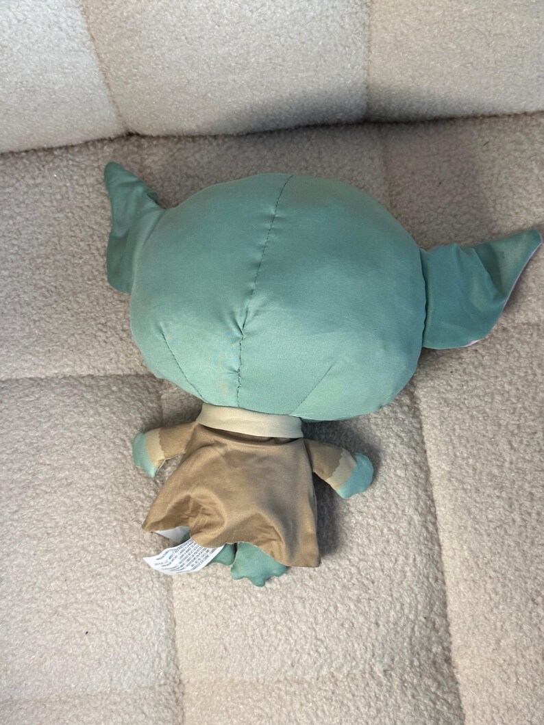 May include: A plush doll of a character with large ears, a green head, and a tan robe. The doll has small arms and feet with blue accents. The doll is lying on a light-colored, textured surface.
