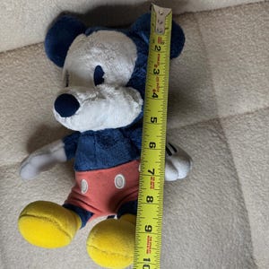 May include: A plush Mickey Mouse toy, approximately 25 cm tall, features a navy blue body, ears, and nose, with a white face and hands. It wears red shorts and yellow shoes. The toy is positioned next to a yellow measuring tape.