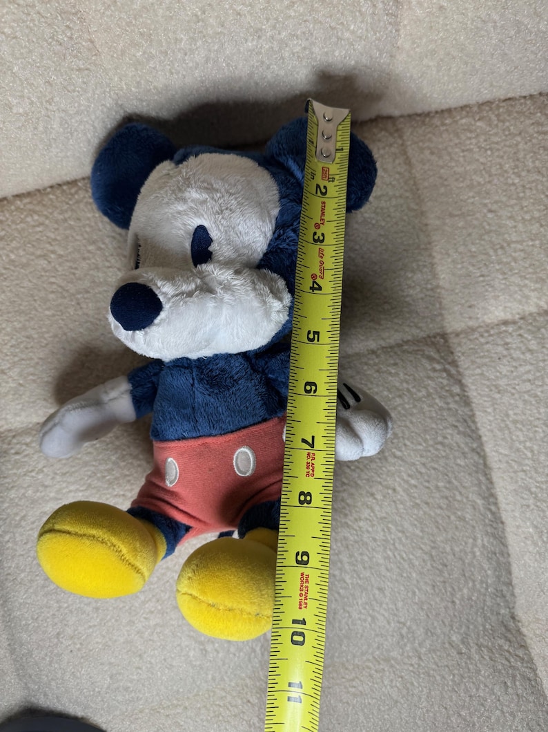 May include: A plush Mickey Mouse toy, approximately 25 cm tall, features a navy blue body, ears, and nose, with a white face and hands. It wears red shorts and yellow shoes. The toy is positioned next to a yellow measuring tape.