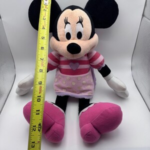 May include: A Minnie Mouse plush doll, approximately 14 inches tall, with black ears, a pink and white striped top, and pink polka-dotted skirt. The doll has large pink shoes and white gloves.