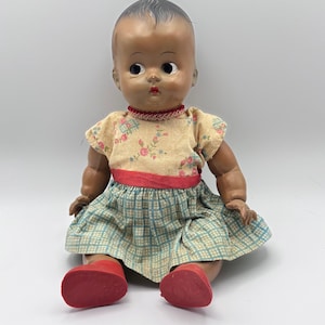 May include: Vintage baby doll with dark skin, wearing a floral print top, a plaid skirt, and red shoes. The doll has a painted face with large eyes and a red mouth. The doll is sitting down.