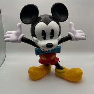 以下が含まれることがあります： 黒い耳、白い顔、青い蝶ネクタイをしたミッキーマウスのフィギュア。赤いショートパンツと黄色の靴を履き、白い手袋をしています。腕は伸ばされ、親指は上を向いています。