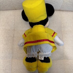 May include: A plush toy of a character wearing a yellow uniform with red trim, a matching hat, and black ears. The character's white gloves and blue pants with lace trim are visible. The toy's yellow shoes complete the outfit.