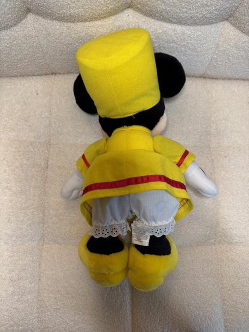 May include: A plush toy of a character wearing a yellow uniform with red trim, a matching hat, and black ears. The character's white gloves and blue pants with lace trim are visible. The toy's yellow shoes complete the outfit.