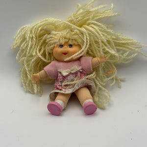 May include: A vintage doll with a soft vinyl face, blonde yarn hair, and a pink dress with a floral print. The doll has pink shoes and is in a seated position. A classic toy.