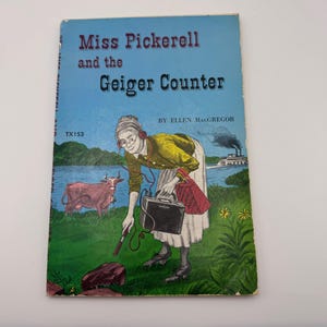May include: A vintage book cover titled "Miss Pickerell and the Geiger Counter" with a blue background. The cover illustration depicts a woman with headphones and a Geiger counter, a cow, and a river with a steamboat. The book is by Ellen MacGregor.