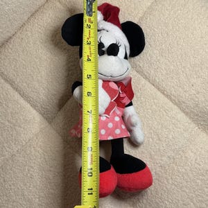 Op de afbeelding: Een Minnie Mouse pluche speelgoed, ongeveer 25 cm hoog, draagt een rood-witte polkadot jurk, een rode strik en een kerstmuts. Het speelgoed heeft zwarte oren, een wit gezicht en rode schoenen. Een gele rolmaat ligt naast het speelgoed.