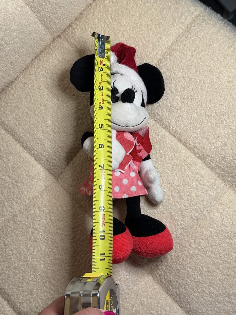 Op de afbeelding: Een Minnie Mouse pluche speelgoed, ongeveer 25 cm hoog, draagt een rood-witte polkadot jurk, een rode strik en een kerstmuts. Het speelgoed heeft zwarte oren, een wit gezicht en rode schoenen. Een gele rolmaat ligt naast het speelgoed.