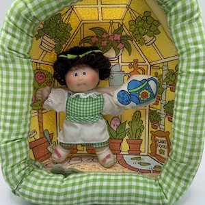 May include: A vintage Cabbage Patch Kids doll in a greenhouse-themed display. The doll wears a green and white checkered dress and holds a watering can. The display has a green and white gingham border and a yellow background with potted plants.