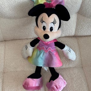 May include: A plush Minnie Mouse doll with a rainbow-colored dress and bow. The doll has black ears, a pink bow, and pink shoes. The dress transitions from yellow to pink, with a pink collar and white gloves.