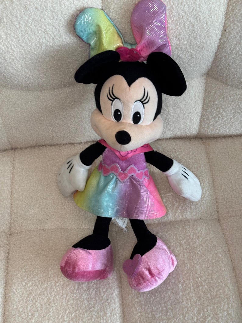 May include: A plush Minnie Mouse doll with a rainbow-colored dress and bow. The doll has black ears, a pink bow, and pink shoes. The dress transitions from yellow to pink, with a pink collar and white gloves.