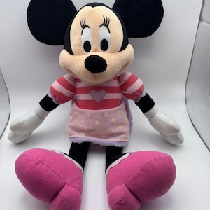May include: A plush Minnie Mouse doll with large black ears, a pink and white striped top, and a pink skirt with gold polka dots. The doll has large pink shoes and white gloves.
