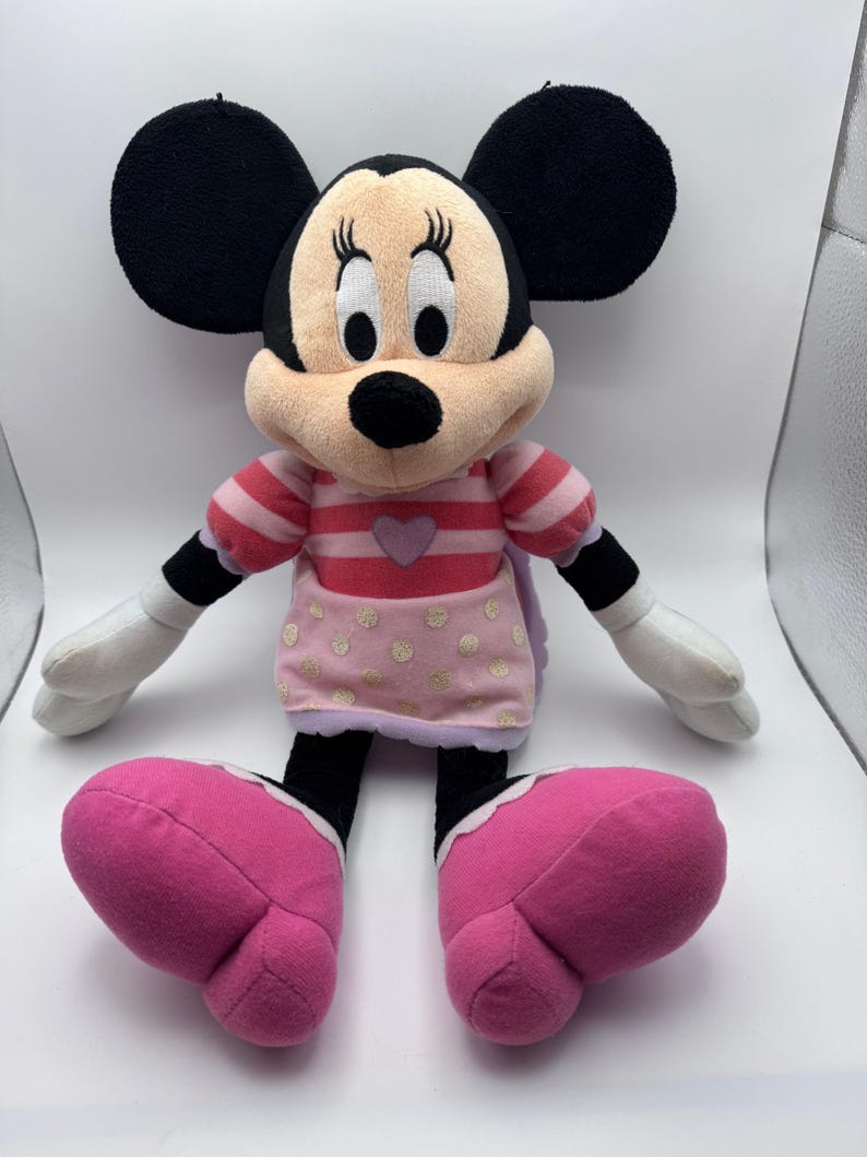 May include: A plush Minnie Mouse doll with large black ears, a pink and white striped top, and a pink skirt with gold polka dots. The doll has large pink shoes and white gloves.